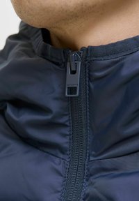 Jūrų laivyno zip-up striukė su aukštu apykakle, lygiu paviršiumi, priekyje su užtrauktuku ir prekiniame ženkle su trauklėmis bei siūlėmis šalia apykaklės.