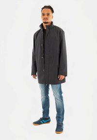 Schwarzer langer Mantel mit hohem Kragen, vorne mit Knöpfen und Seitentaschen, getragen über einem schwarzen Hemd und blauen Jeans, kombiniert mit blauen Turnschuhen.