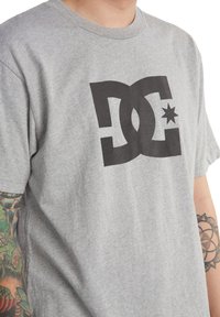 Šedé bavlněné tričko s velkým černým logem DC a hvězdným akcentem, s kulatým výstřihem a krátkými rukávy.