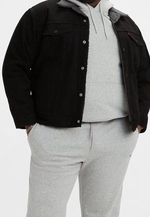 Man met een zwarte jas over een lichtgrijze hoodie en bijpassende lichtgrijze sweatpants, handen in de zakken, staande tegen een witte achtergrond.