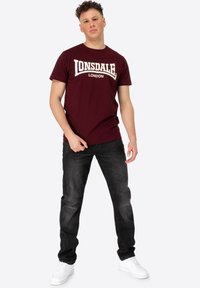 Burgundi pamut póló fehér "LONSDALE LONDON" grafikával, sötét farmernadrággal és fehér tornacipővel párosítva, kerek nyakkivágással.