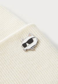 Tessuto bianco a coste con una spilla argentata raffigurante un gatto con occhiali da sole neri, che presenta un design facciale dettagliato e piccole minuterie decorativi.