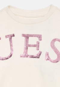 Κοντινή λήψη του λαιμού ενός κρεμ φανελιού με μεγάλα ροζ στρας γράμματα που σχηματίζουν μέρος μιας λέξης.