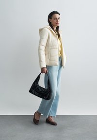 Φουσκωτό μπεζ μπουφάν με κουκούλα, κίτρινη μπλούζα, ανοιχτό μπλε παντελόνι με φαρδιά πόδια και σκούρα καφέ παπούτσια. Κρατάει μια μαύρη λουστρινένια τσάντα.