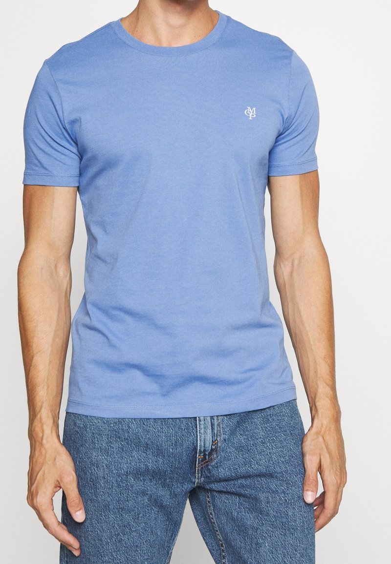 Homme portant un t-shirt ajusté bleu clair à manches courtes avec un petit logo blanc sur la poitrine et un jean bleu, debout devant un fond uni.