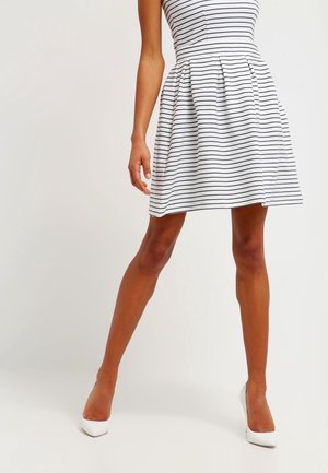 Femme portant une robe blanche sans manches avec des rayures horizontales noires et des chaussures blanches à talons, debout devant un fond uni.