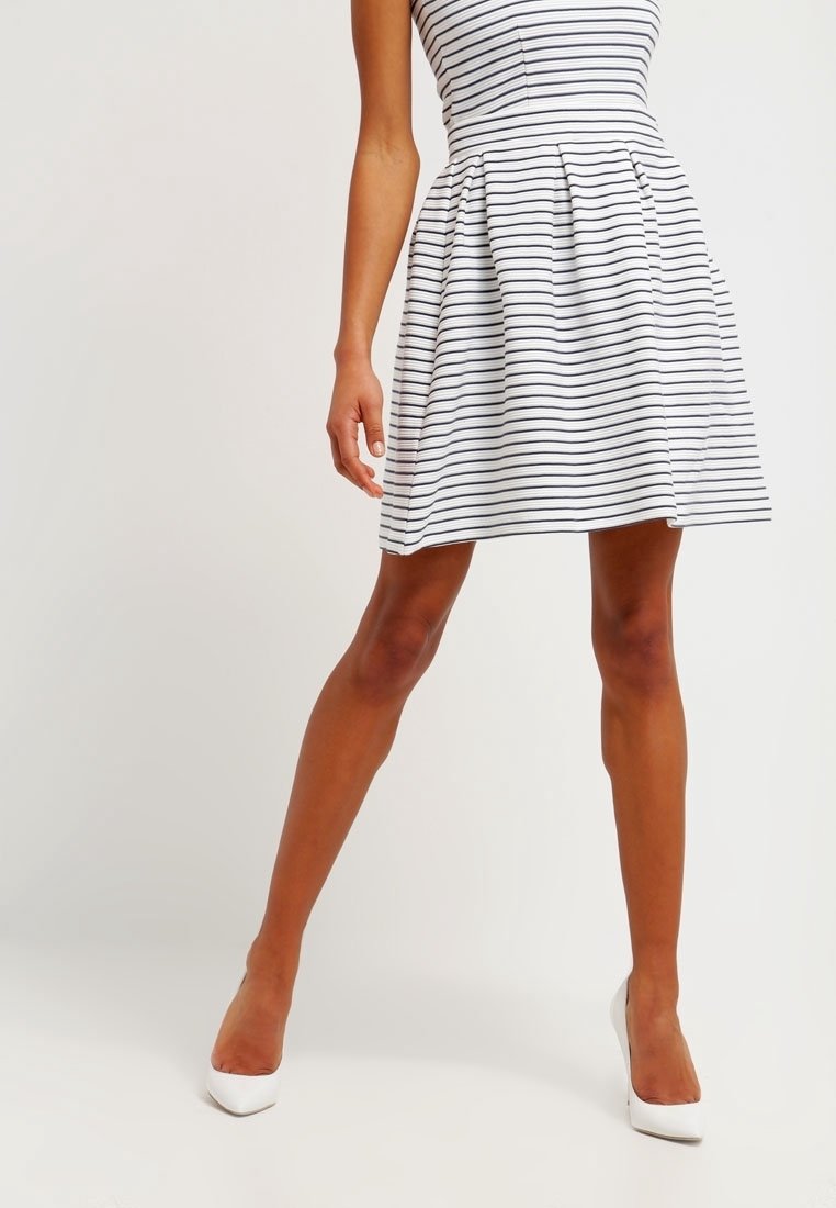 Femme portant une robe blanche sans manches avec des rayures horizontales noires et des chaussures blanches à talons, debout devant un fond uni.
