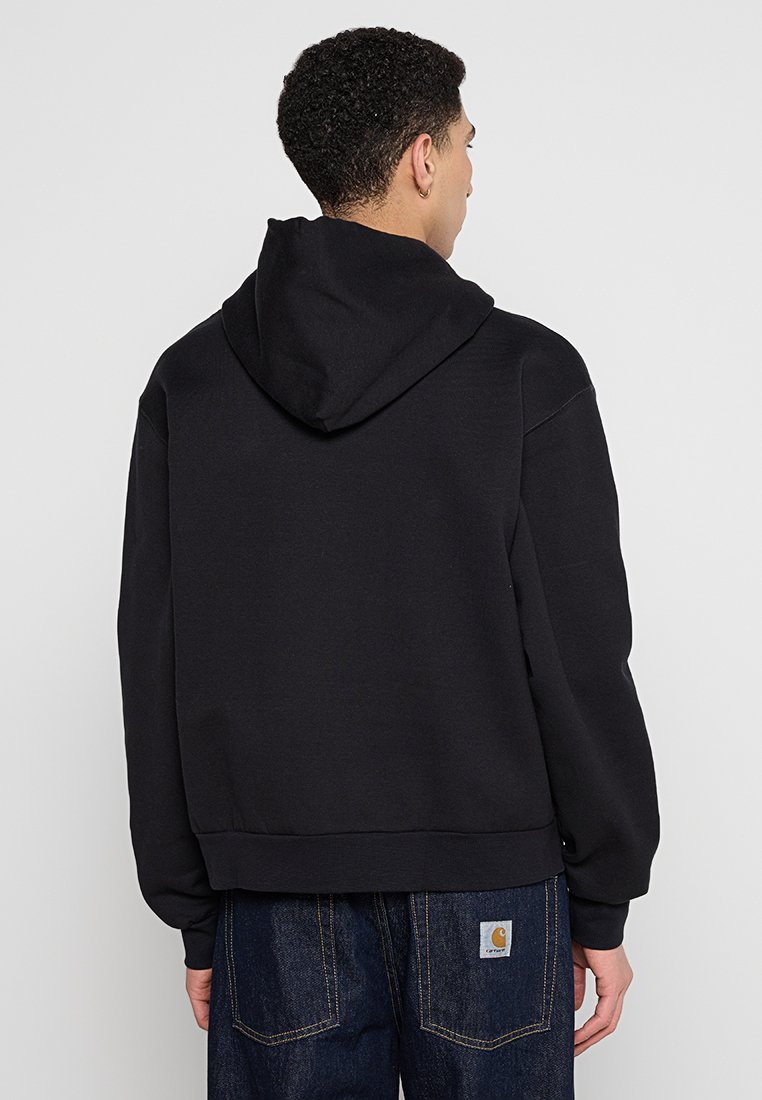 Hoodie noir avec une capuche à cordon, des poignets et un ourlet côtelés, présentant une coupe décontractée et une texture lisse, associé à un jean foncé.