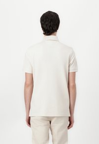 Beige polo met korte mouwen, klassieke kraag en een textuurafwerking. Draagt met lichtbeige broek, gezien vanuit de achterkant tegen een witte achtergrond.