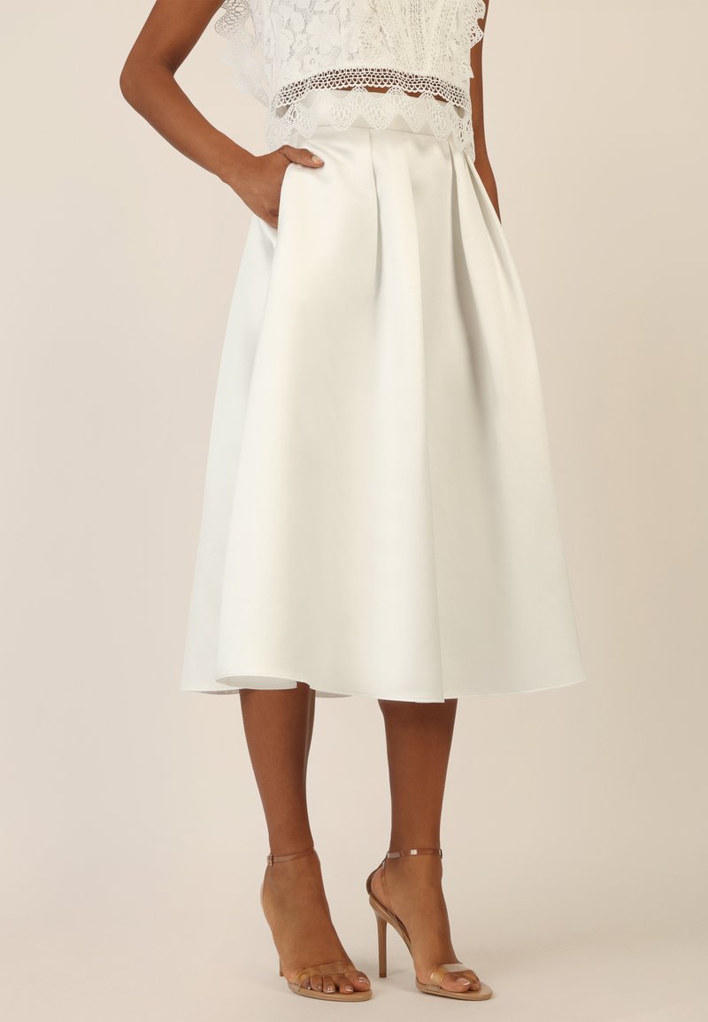 Una gonna bianca, lunga fino al ginocchio, realizzata in tessuto liscio con plissettature e tasche laterali, abbinata a sandali con tacco nude.