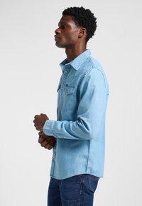 Jeune homme avec une coupe afro courte, portant une chemise en jean bleu clair et un jean bleu foncé, debout de profil les mains jointes.