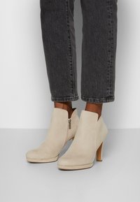 Beige Wildleder-Stiefeletten mit einem seitlichen Reißverschluss, einer schmal zulaufenden Form und einem hohen Absatz. Kombiniert mit dunklen, verkürzten Jeans. Glatte Textur.