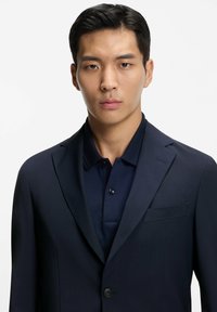 Granatowa marynarka z klapami z wycięciem, zapinana na jeden guzik, z kieszenią na piersi, noszona na pasującej ciemnej koszulce polo. Gładka faktura i dopasowany krój.