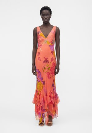 Femme portant une robe corail sans manches, longue jusqu'au sol, avec des motifs floraux violets, jaunes et roses, et un ourlet volanté, se tenant devant un fond blanc.