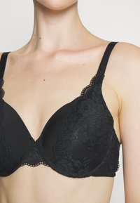 Black lace bra with smooth cups, scalloped edges, and adjustable straps. Features floral pattern on the lace overlay.