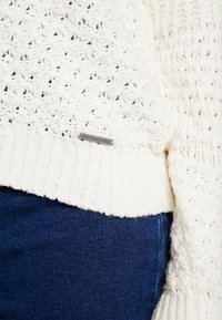 Biała sweterkowa bluzka z teksturowanym wzorem i ściągaczem u dołu, z małą metalową metką logo na dole. Zestawiona z ciemnymi dżinsami.