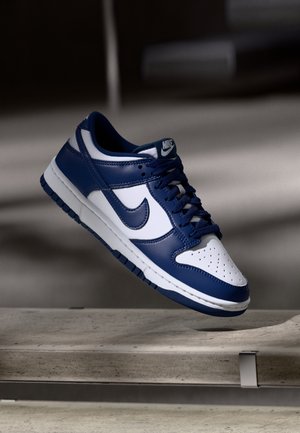 Blå og hvide Nike lavsko med snørebånd, der svæver over en betonbænk i et dunkelt oplyst miljø.