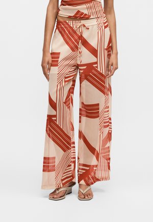 Spodnie o szerokich nogawkach z czerwonymi geometrycznymi paskami na beżowej tkaninie, noszone z dopasowanym topem i beżowymi sandałami z paseczkami między palcami.