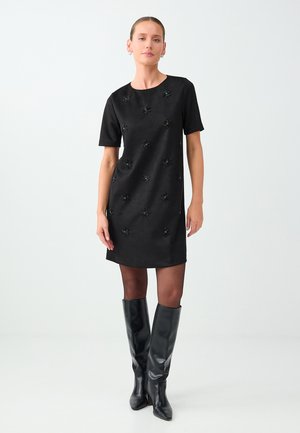 Une femme porte une robe noire à manches courtes ornée de motifs floraux et des bottes noires montant jusqu’aux genoux, sur un fond de studio clair.