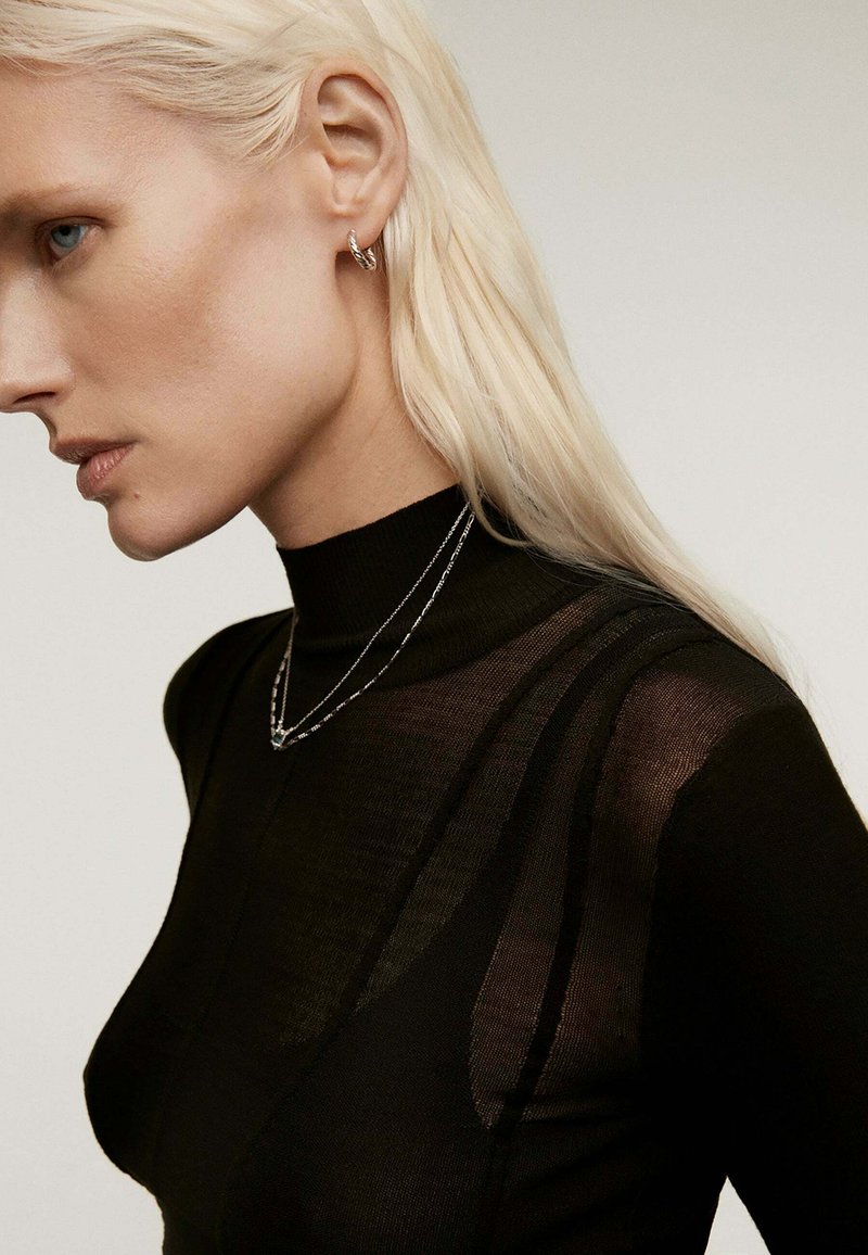 Top negro de cuello alto con detalles transparentes, combinado con cadenas de plata en capas y un pendiente de aro retorcido. Textura suave con un silueta ajustada.