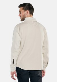 Beige Langarmhemd mit strukturierter Oberfläche, mit Kragen und seitlichen Schlitzen an den Manschetten. Rückansicht mit einem Logo in der Nähe des Ausschnitts.