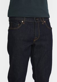 Jean en denim bleu foncé avec coutures orange, poches avant, bouton et passants pour ceinture, porté avec une chemise sombre rentrée dedans.