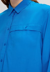Blauwe shirt van gladde stof, met een knopsluiting kraag en een enkele borstzak met een knoop. Naaddetails zichtbaar.