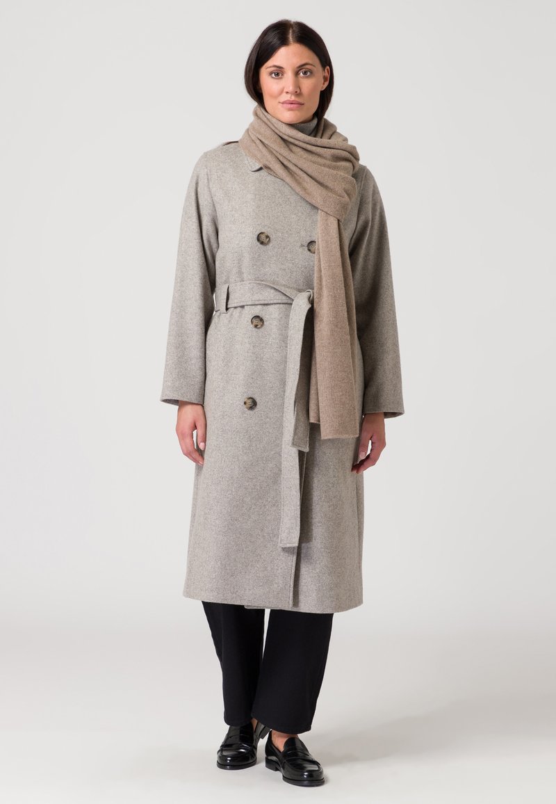 Cappotto in lana grigio con bottoni doppi e una cintura, abbinato a una sciarpa beige. Pantaloni neri a gamba larga e scarpe nere lucide completano il look.