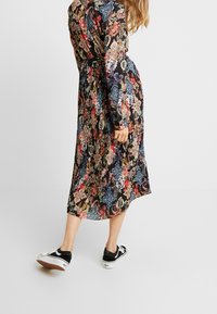 Schwarz langärmliges Kleid mit bunten Blumenmustern, gefaltetem Design, Taillenknoten und kombiniert mit schwarzen Sneakers.