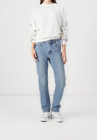 Sudadera blanca con bordados florales en las mangas, combinada con jeans azul claro. La modelo lleva zapatillas blancas. Diseño sencillo y ajustado.