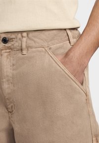 Man som bär beiga byxor med handen i framsidan av fickan, vilket visar knappen, bältesöglor och tygtextur på nära håll.