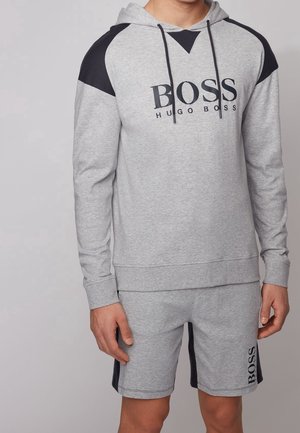 Homme portant un sweat à capuche gris avec des panneaux noirs aux épaules et un short gris assorti avec des bandes noires sur les côtés, tous deux ornés du logo BOSS.