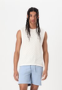 Lys cremet ærmeløs strikbluse med diamantm�ønster, parret med lyseblå shorts. Stoffet ser blødt og let tekstureret ud.