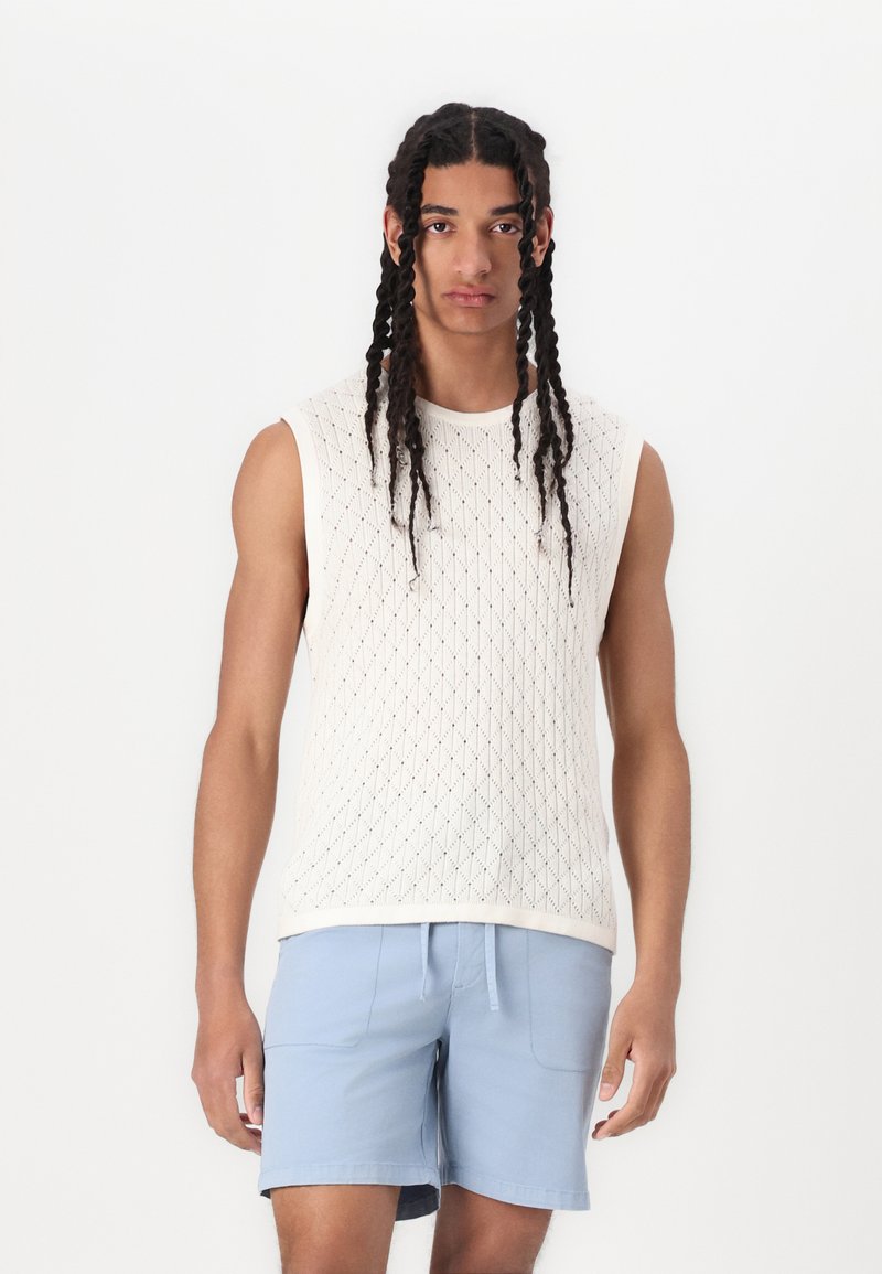 Top sans manches en tricot crème clair avec un motif en diamant, associé à un short bleu clair. Le tissu semble doux et légèrement texturé.