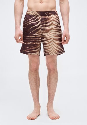 Mees, kes kannab pruuni ja beeži tiigritriibulisi lühemaid pükse, seisab paljajalu tavalise valge taustaga.