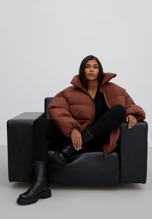 Bruine pufferjack met hoge kraag, zwarte, strakke trui en chunky zwarte veterschoenen, zittend op een zwarte fauteuil.