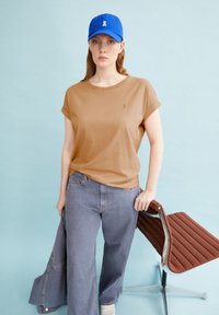 Braunes T-Shirt mit kurzen Ärmeln, hellblaue Jeans und eine blaue Mütze mit einem weißen Logo. Modell hält einen braunen gepolsterten Stuhl. Hellblauer Hintergrund.