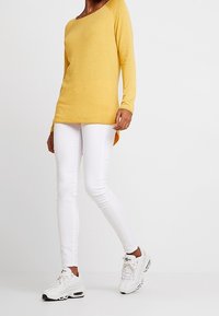 Top giallo a maniche lunghe, dalla vestibilità ampia, abbinato a jeans bianchi aderenti e scarpe da ginnastica bianche. Tessuto morbido, scollo rotondo e orlo a lunghezza variabile.