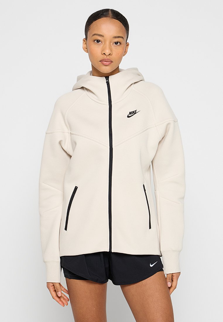 Cremefarbener Nike-Hoodie mit Reißverschluss, schwarzen Akzenten, Fronttaschen und lockerer Passform. Verfügt über eine Kapuze und strukturierten Stoff.