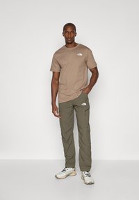 Brun t-skjorte med logo, grønne cargo-bukser med lommer og hvite joggesko med mørke snørebånd, stående på en ensfarget bakgrunn.