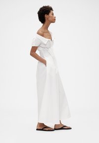 Witte off-shoulder jurk van katoen, met een knopenrij aan de voorkant, korte mouwen en twee zijzakken. Gecombineerd met zwarte slippers.