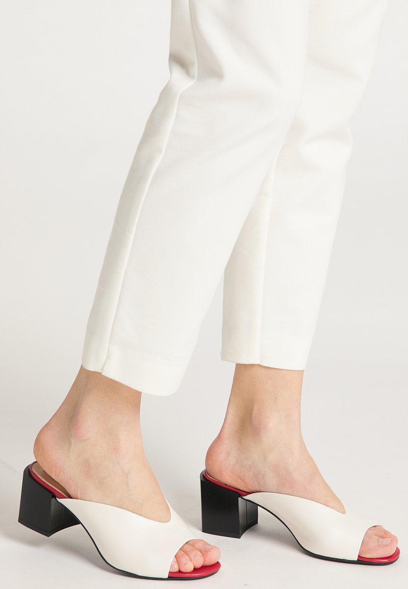 Witte open teen mules met een V-vormig ontwerp, voorzien van een chunky zwarte hak en een rode accent op de inlegzool. Gladde leren textuur.