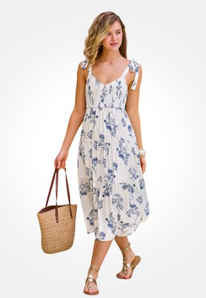 Žena v bielych midi šatách s modrým kvetinovým vzorom a viazanými ramienkami, nesie pletenú tašku a má na sebe sandále s remienkami.
