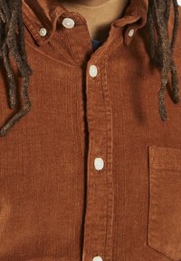 Camisa de pana color óxido con diseño abotonado, botones blancos y un bolsillo en el pecho, mostrando una textura acanalada.