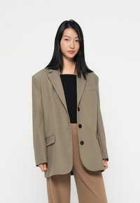Overmaatse bruine blazer met scherpe revers, twee voorkeurs en drie zwarte knopen. Draagt over een zwarte top en beige broek.