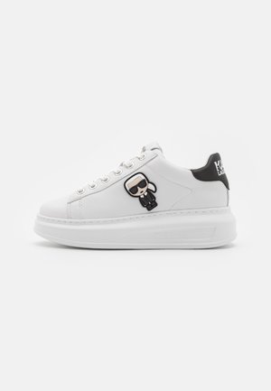 White low-top sneaker with thick sole, metal eyelets, and a small cartoon figure wearing sunglasses on the side; black heel tab with white text.