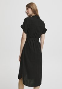 Rochie neagră dintr-un material ușor, cu mâneci scurte răsucite, o curea legată în talie și o fante laterală pentru ușurința mișcării.