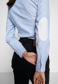 Camicia azzurro chiaro con maniche lunghe, caratterizzata da toppe bianche sui gomiti con un piccolo motivo a cuori neri. I pantaloni neri sono parzialmente visibili.