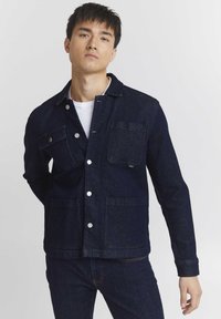 Dark denim jacket with a button-up design, two front pockets, and contrasting stitching, paired with a white T-shirt underneath.