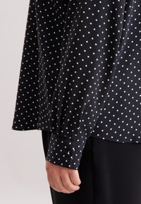 Zwarte blouse met witte stippen, zachte stof, lange mouwen, knoopmanchetten en een ontspannen pasvorm.